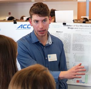 A young man stands in front of an academic poster, gesturing towards it as he speaks.