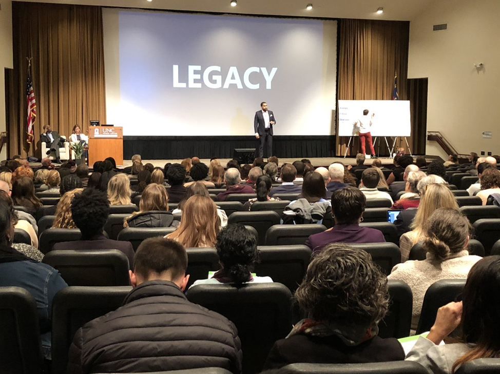 A man speaks to a crowded auditorium. Behind him, a large screen displays the word "legacy." Near him onstage, a person facing a white poster is transcribing the speaker's ideas into art.
