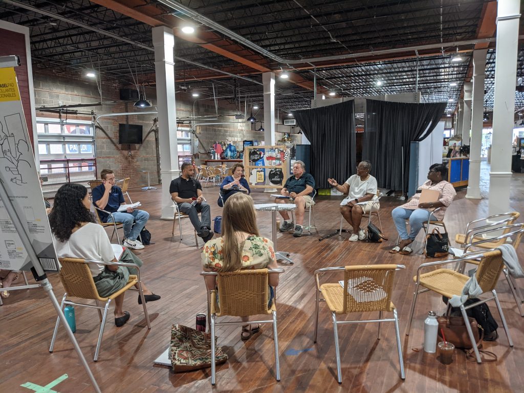 A group of people sit in chairs in a circle, inside a large room that looks like a warehouse.