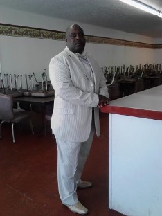 A Black man in a white suit stand and looks at the camera