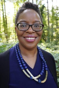 Spurlock has short hair and dark stylistic glasses and pink lipstick. Her blazer is black and her shirt underneath is indigo blue. She has two necklaces with prominent dark blue beads and gold-color bars. She is standing in front of trees.