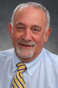 Strauss has a neutral gray background behind him. He has a clean beard and mustache and shorter gray hair. His shirt is white and light blue stripes. His tie has yellow and blue stripes.