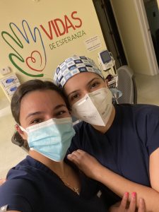 Two dental workers pose for a selfie. Both are wearing dark blue scrubs and surgical masks. The person on the left is wearing a UNC branded bandana. Behind them on the wall is a stylistic hand that swirls into a heart shape on the palm. Next to it are the words vidas de esperanza.