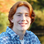 Troy has long straight reddish hair and a white shirt with blue stripes. Fall trees are in the background.