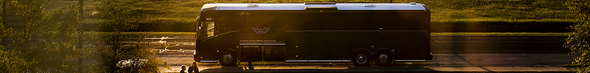 A tightly cropped image of the Tar Heel Bus Tour bus parked near a large field. The rising sun provides warm oranges and yellows throughout the image.
