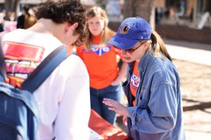 Someone wearing a blue Durham bulls cap, denim jacket and orange You Can Vote shirt talks to a student about the voter registration form. The students are outside of the union at UNC-Chapel Hill