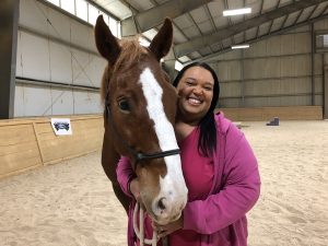 April Parker is wearing a magenta jacket and standing with a brown and white horse in a large enclosed facility filled with sand.