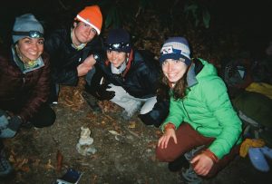 Four students squat on the ground in front of a statue of six rocks stacked on each other. Each of the students wear jackets, beanies and head lamps. The photo was taken past sun set and uses artificial light from the camera or other source.