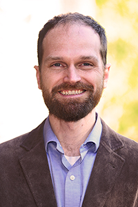 Ryan Nilsen has short brown hair and a full beard, mustache and goatee. The facial is brownish-red. Ryan wears a blue shirt and brown corduroy blazer. The background is blown out from the sun but some golden leaves are visible.