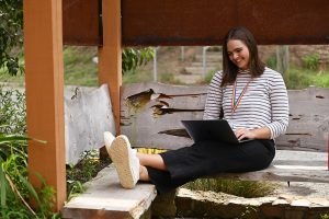 Sarah wears a black and white stripe shirt. She sits on an l shaped bench under a cover while looking down at a black laptop. She has her feet propped on the joining bench.